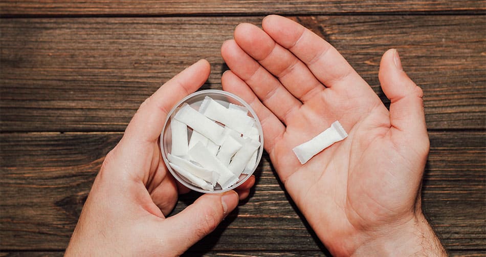 Man holding a pack of nicotine pouches in one hand with another placed in their palm on top of a wooden table.