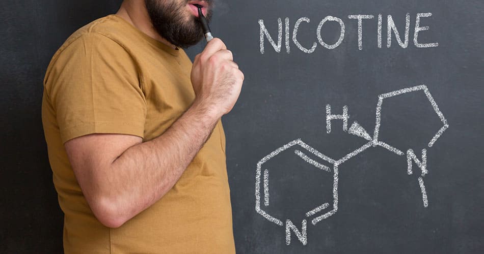 Man vaping on an e-cigarette with nicotine written on a chalkboard in the background.