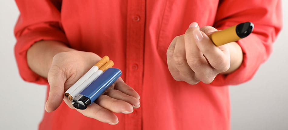 Person holding cigarettes and a lighter in one hand and a disposable vape in the other hand