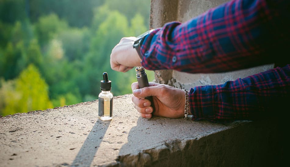 Man filling vape kit with e-liquid bottle dripper on a concrete wall.