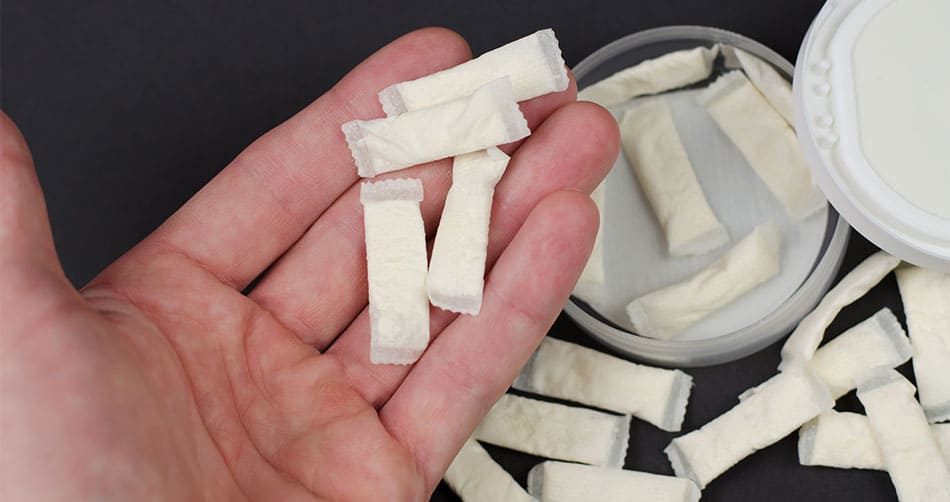 Empty tub with nicotine pouches scattered on a table with person holding a pouch close to the camera.