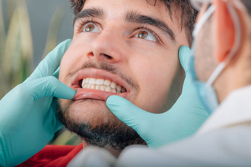 Man looking up with teeth clenched and mouth open, with a dentist pulling down bottom lip and examining.