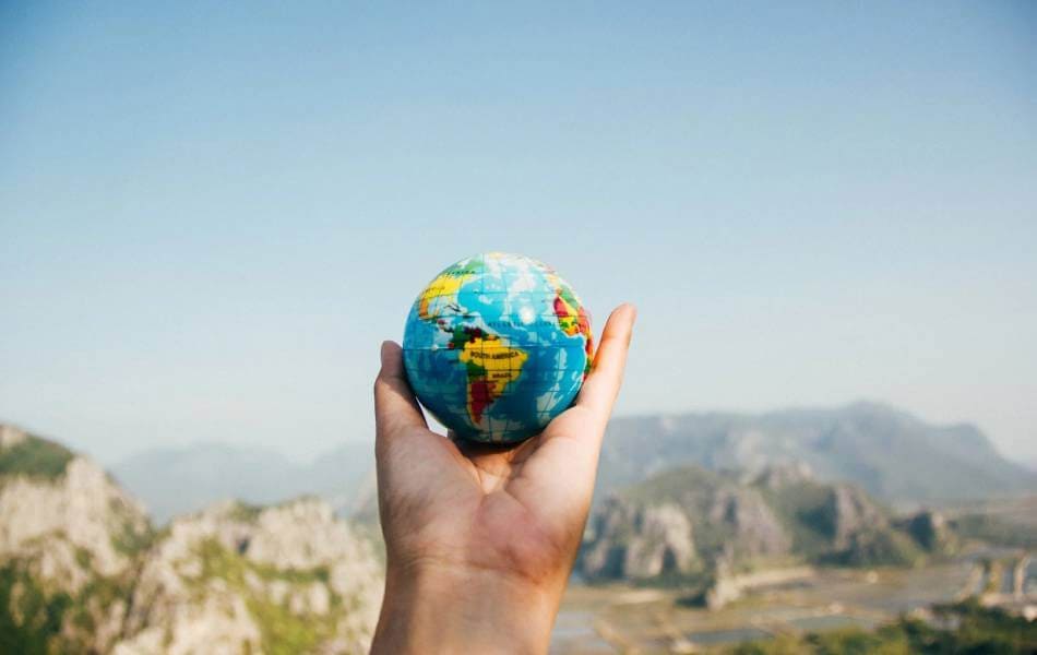 First person photo looking at a stretched out arm with a hand holding a small globe in their hand towards a mountain terrain with a clear blue sky.