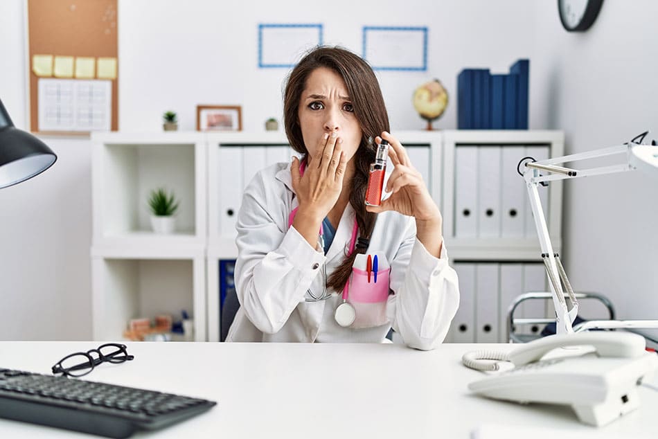 Healthcare professional in white lab coat holding up a vape device in one hand, with their other hand against their mouth with a shocked impression on their face.