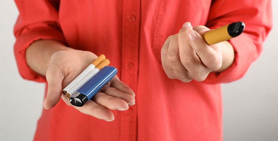 Person holding a vape kit in one hand and cigarettes with a lighter in their other hand.