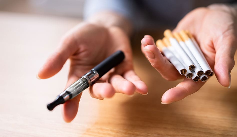 Person holding vape kit in one hand and cigarettes in the other.