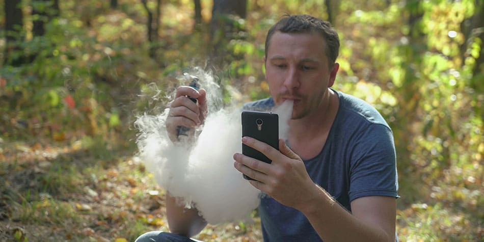 Man sat in woods vaping e-cigarette whilst reading on his phone.
