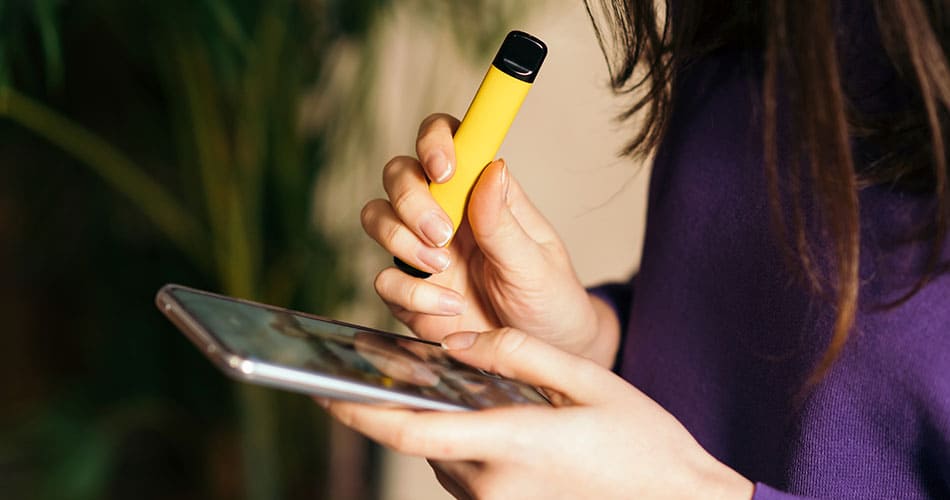 Woman stood indoors vaping a yellow disposable vape looking down on her phone.