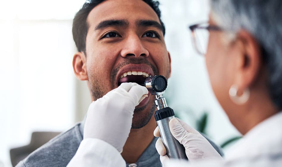 Doctor looking down the throat of a patient with a wooden stick pressed against the tongue and a torch shining.