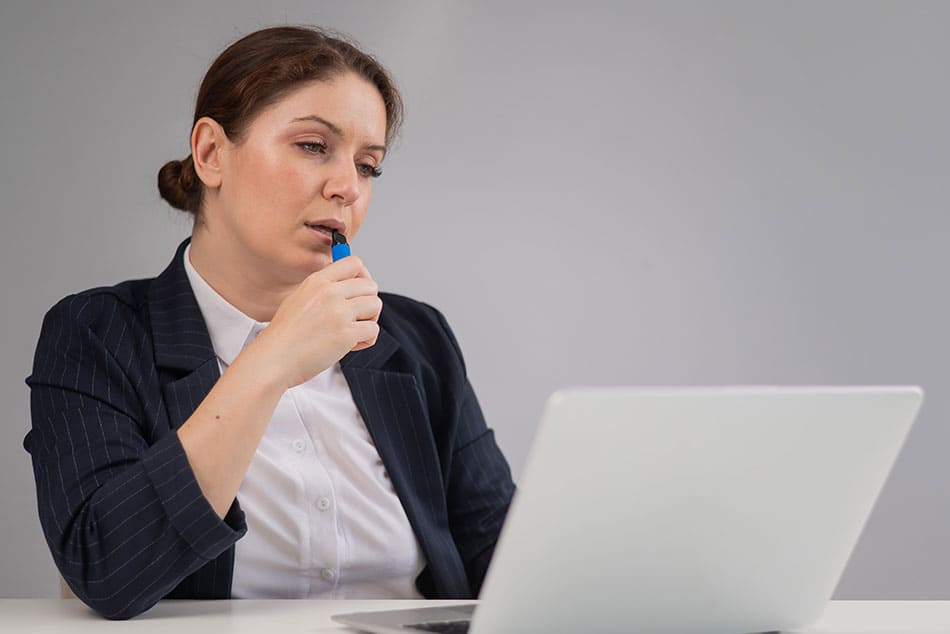 Woman looking at laptop with a disposable vape in her hand pressed against here lips.