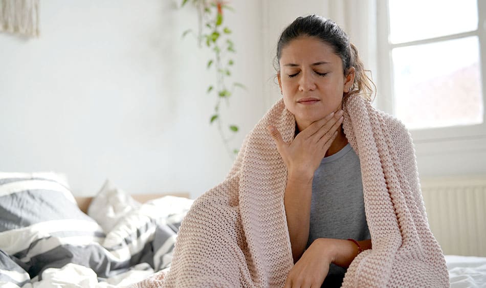 Woman sat on bed with a blanket wrapped around her holding her throat with a painful look on her face.