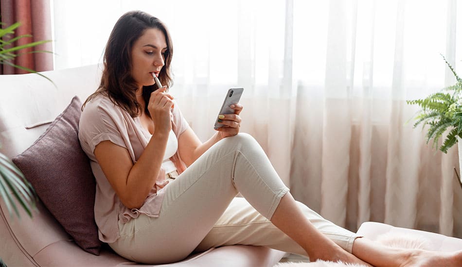 Woman sat on chair looking at her phone whilst vaping an e-cigarette.