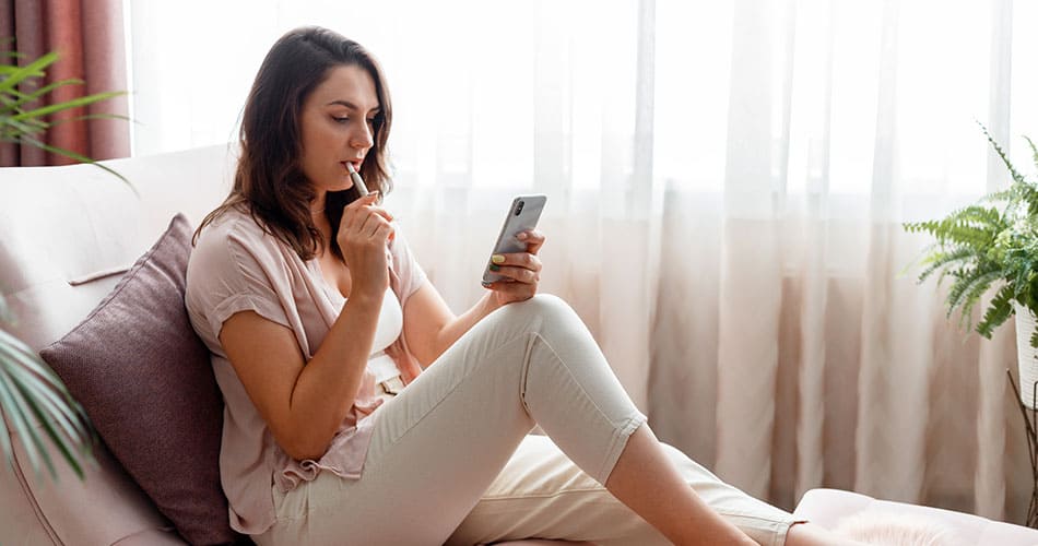 Woman sat inside on a sofa next to a window vaping an e-cigarette and looking at her phone.