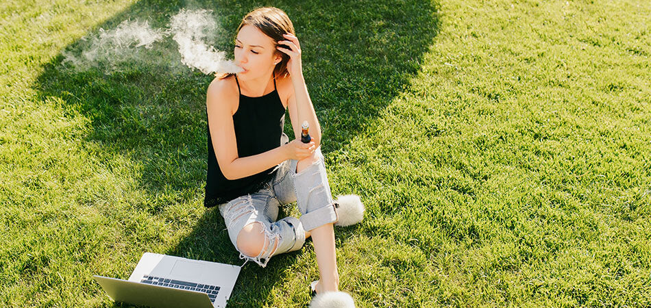 Woman sat on green grass whilst vaping next to laptop.