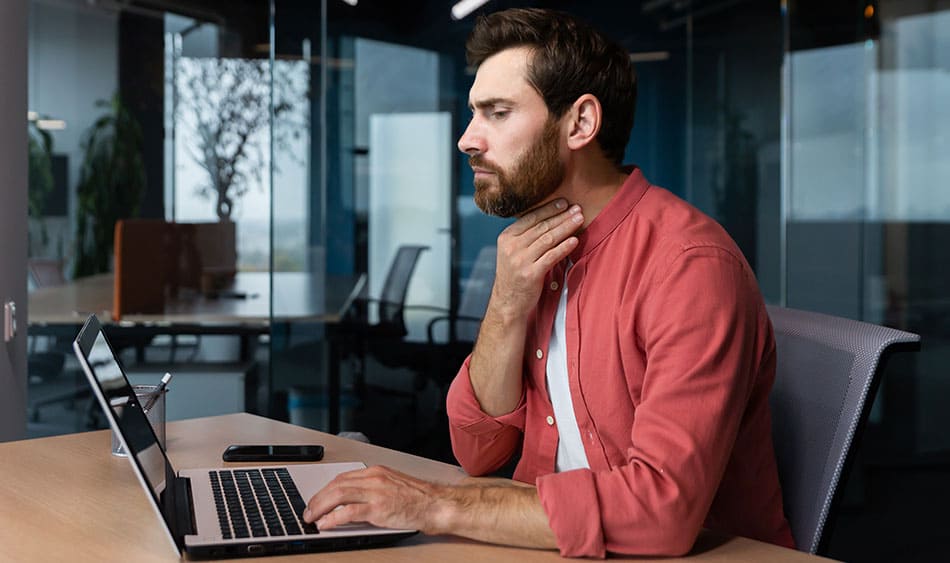 Man sat at table holding his throat looking towards laptop screen.