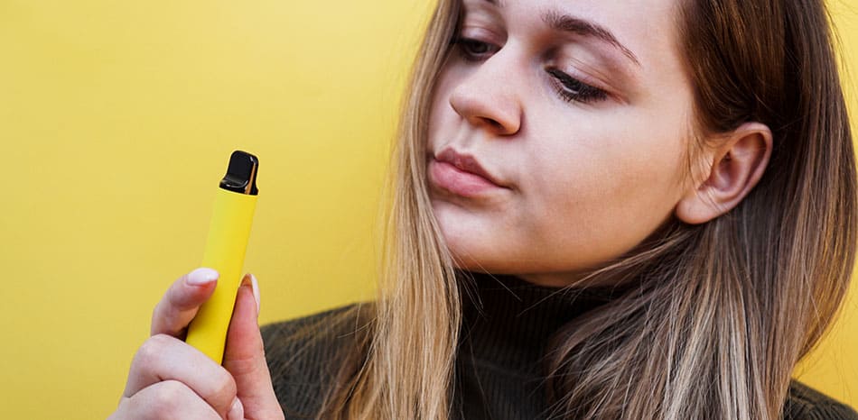 Woman stood in front of yellow background looking at a yellow disposable vape device