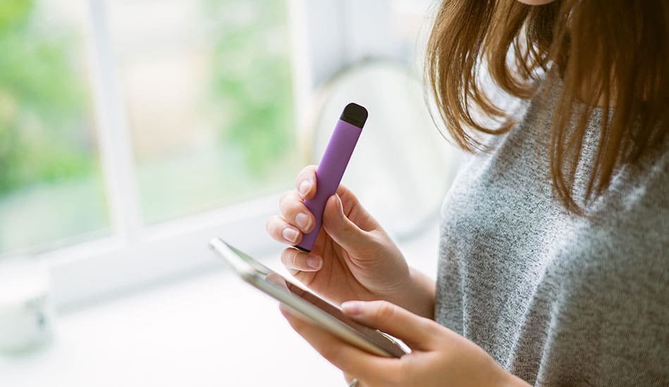 Woman looking down at her phone while holding a purple disposable vape in her hand.