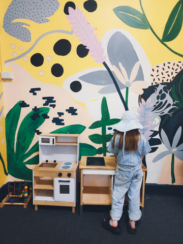 Multi coloured mural with young girl wearing blue hat playing at a toy kitchen