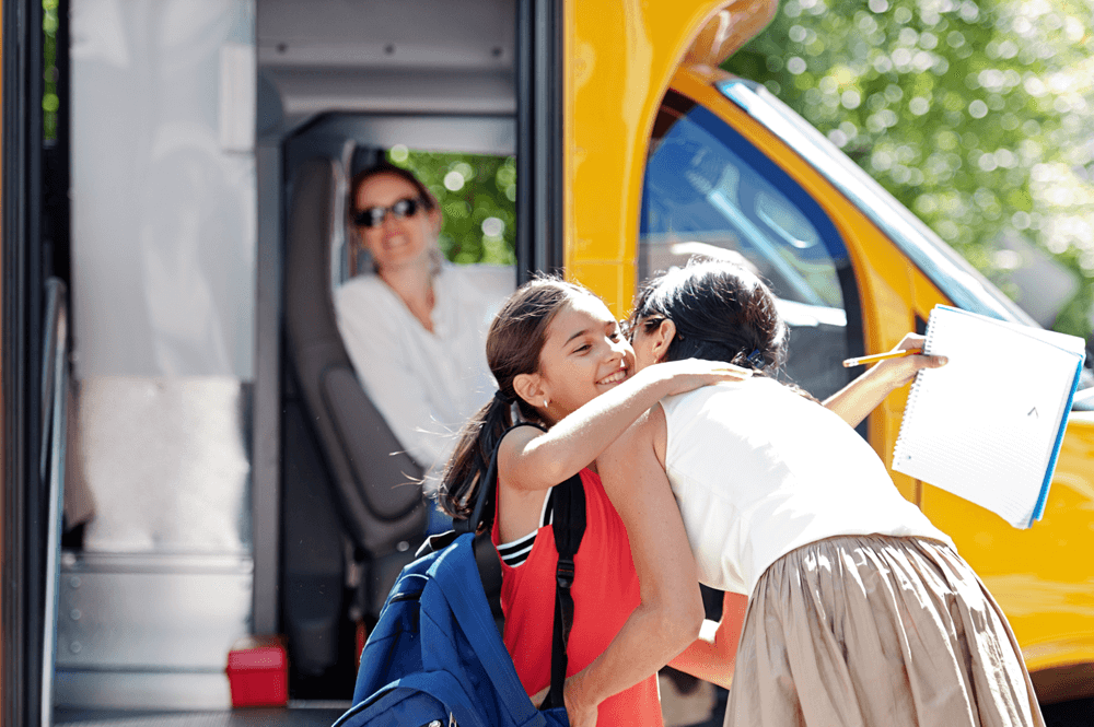 girl in red shirt hugging a woman with a school bus in the background