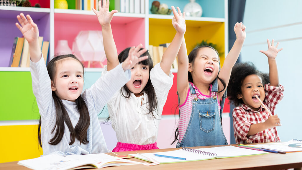 four young children sitting at a desk and raising their hands in the air