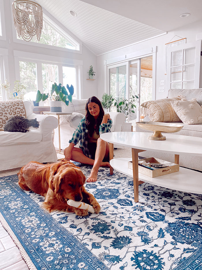 Woman and a dog eating a rawhide sit on a floral area rug in a mostly white living room. 