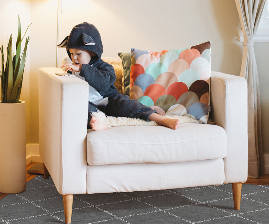 Child sitting on an arm chair on the Medina Moroccan Diamond Grey washable rug.playing with a mobile phone 
