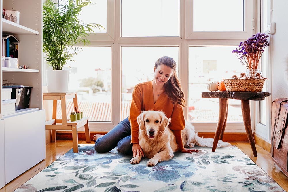 A lady with her dog sitting on the My Magic Carpet Sasha Floral Cream Blue washable rug. 
