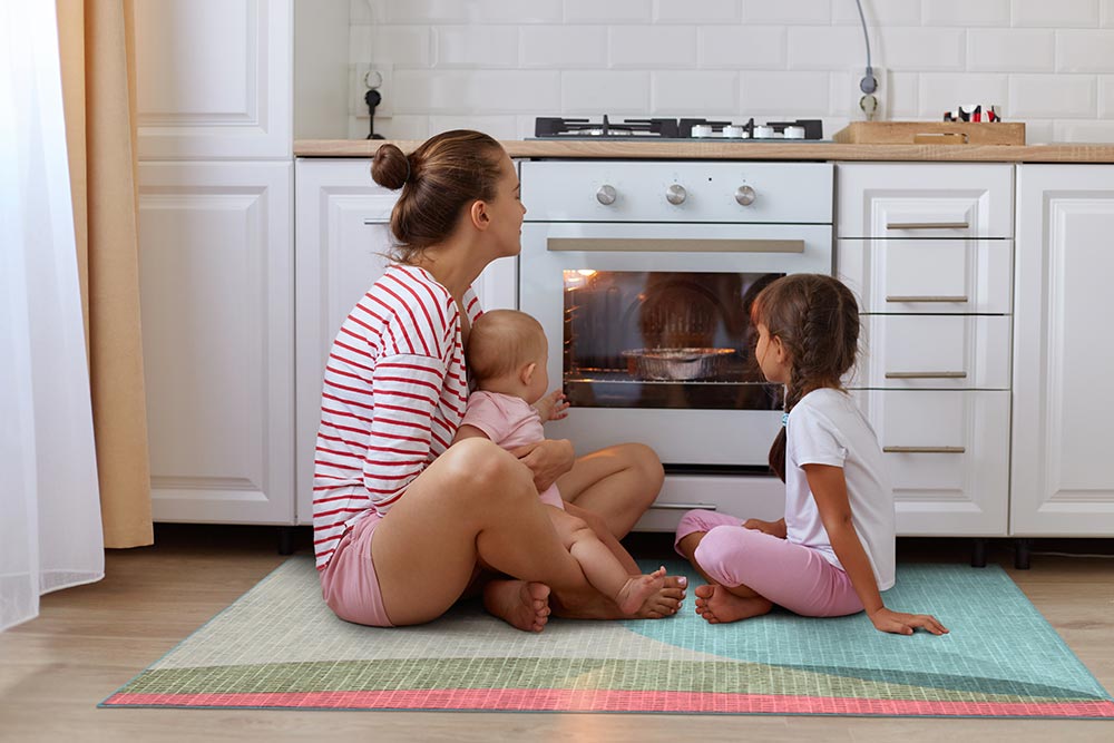 Mom and kids sitting on the kitchen floor in front of an oven on the My Magic Carpet Cove Green washable runner rug.