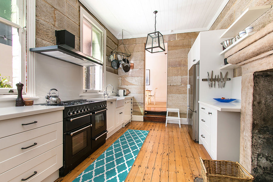 Image of a stone tiled kitchen with white drawers, black appliances, wood floors and a teal runner rug. 