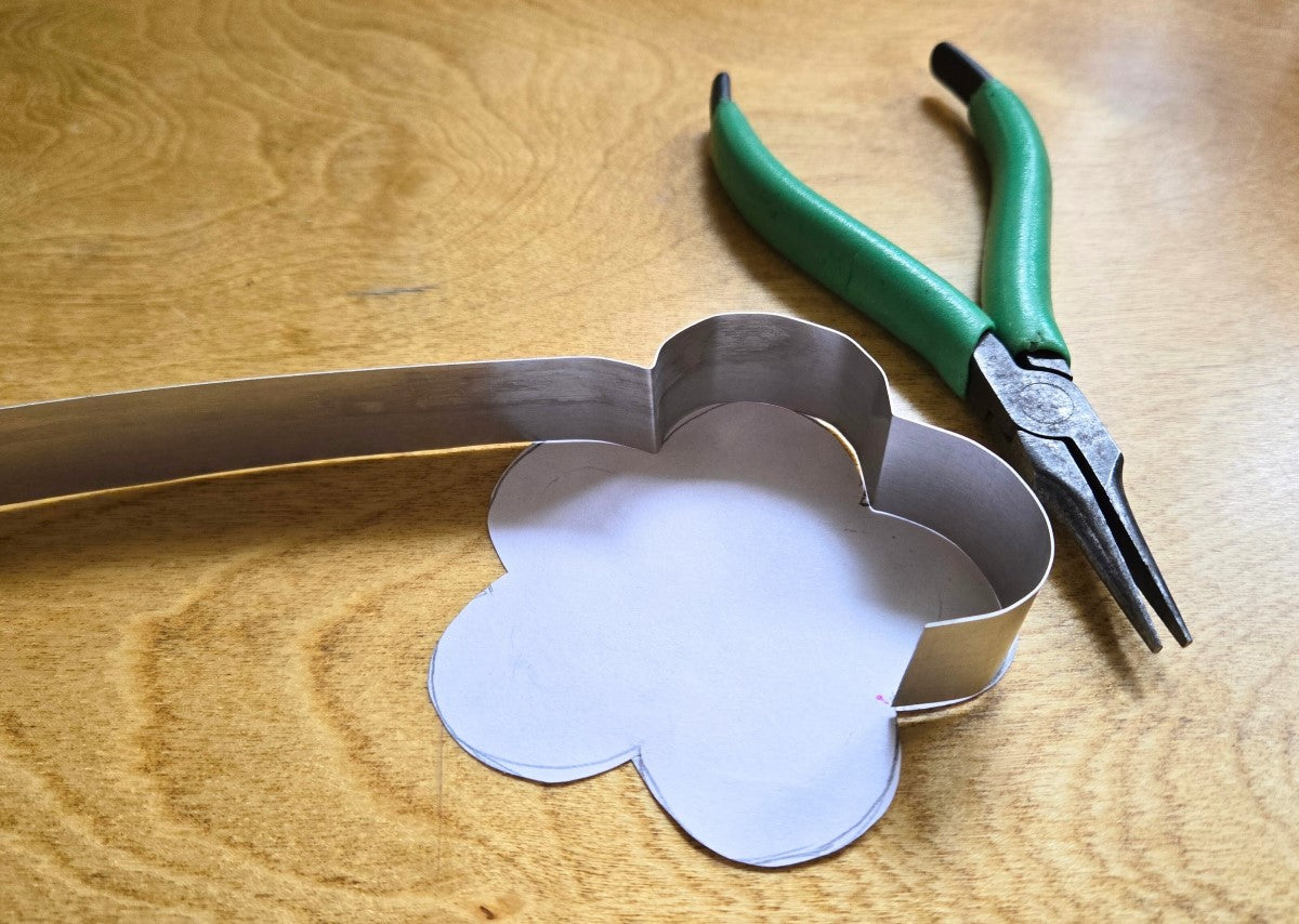 This image shows how Erin makes her own clay cutters. A paper shape lays on the table, a pair of metal shaping pliers lays to the side, and a strip of metal is shown that was bent to follow the outlines of the paper shape.