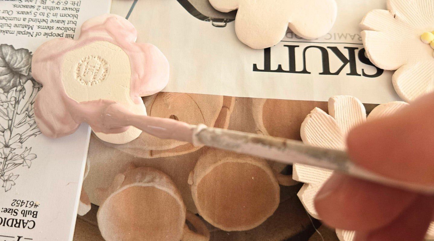 The image shows a hand holding a brush that is brushing pink glaze onto the back of a flower.