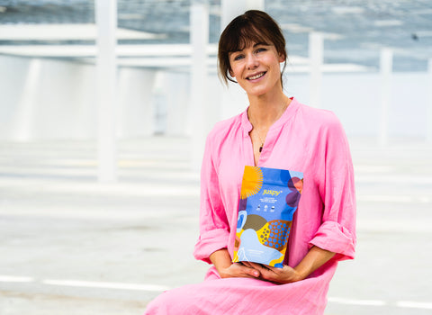Leonie Lynch founder of Juspy holding a pouch of Juspy sitting at water wearing a pink dress