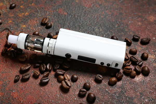 Vape berbentuk kotak berwarna putih dengan tombol hitam diletakkan dekat biji kopi