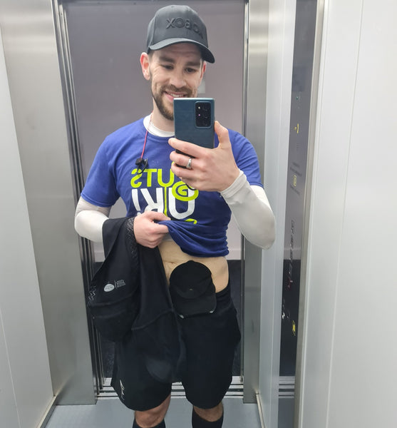 Ant prend un selfie miroir après une course, montrant son sac de stomie et portant son t-shirt Guts UK Charity sur un haut blanc à manches longues, un short de course et une casquette de baseball.