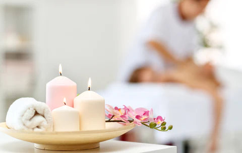 A neutral image with white and pink lit candles in the foreground and a blurred image of a woman receiving a back massage in the background