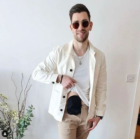 Ant is smiling at the camera and is holding up the bottom of his white t-shirt to reveal a black ostomy bag. He wears a casual white jacket, sunglasses and beige trousers.