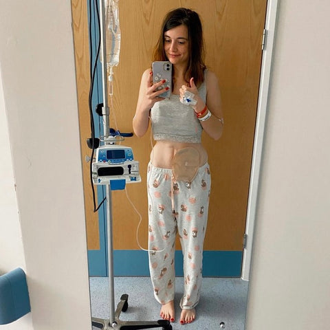Amy est debout, tenant le pouce levé devant un miroir d'hôpital, portant son pyjama, attachée à une perfusion intraveineuse et montrant son sac d'iléostomie.