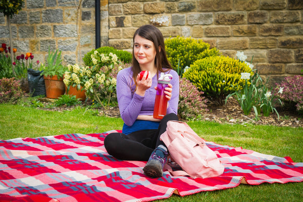 Amy is sat on a red tartan blanket with an apple and a drink in her hands, wearing a purple long-sleeve crop top, black Comfizz leggings and walking boots