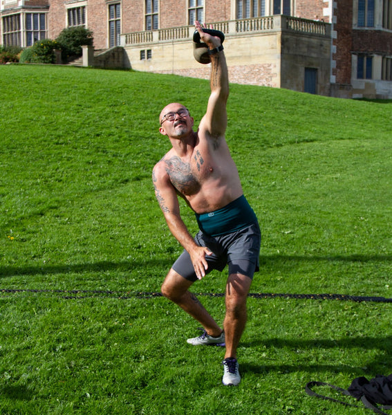 Gareth porte deux de nos ceintures colorées de niveau 1 de 7", ce qui équivaut à un support de niveau 2. Il est accroupi et soulève une kettlebell.