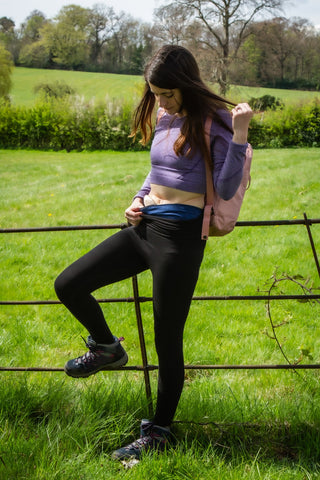 An image showing Amy outside near a fence, having a break on a countryside walk, dressed in Comfizz leggings, a purple long sleeved crop top and showing the top of her ostomy bag and Comfizz ostomy waistband marl in colour indigo