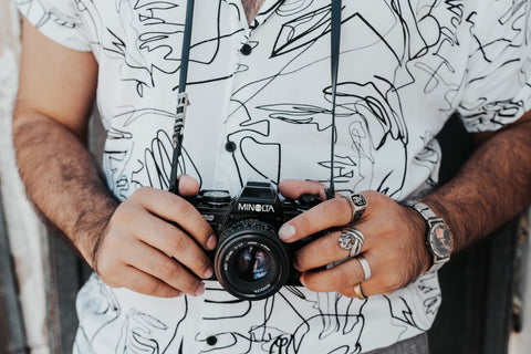 White shirt cropped image from chest to waist with black, scribble print and man's hands with rings on holding a camera