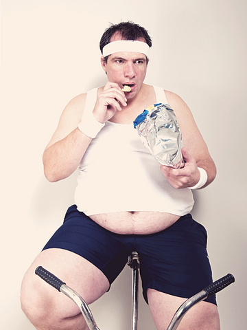 overweight guy on an indoor bicycle eating junkfood.