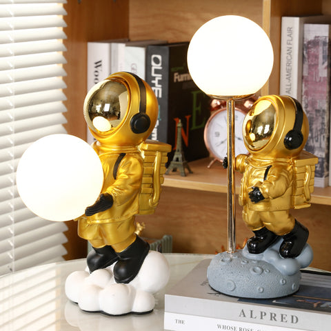 Two astronauts in gold space suit. One holding moon in both hands and another holding to a pole with moon attached on top. Astronaut Moon Lamps by Allthingscurated.
