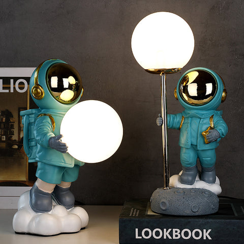 Two astronauts in blue space suit. One holding moon in hand and another holding to a pole with moon attached on top. Astronaut Moon Lamp by Allthingscurated.