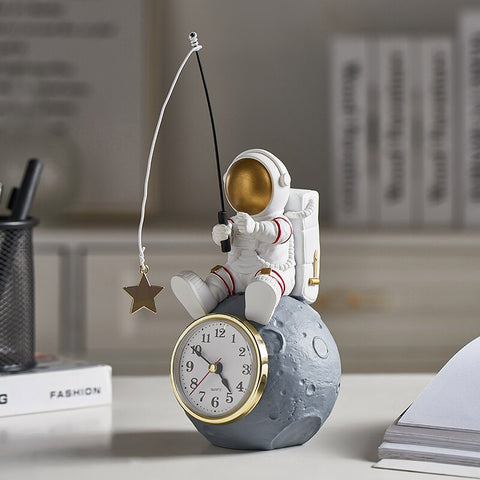 Allthingscurated Desk Clock featuring an astronaut sitting atop a moon rock fishing a star. Astronaut is dressed in a white space suit with a gold space mask, holding a fishing rod with a dangling gold star. Size of clock measuring height 24cm, width 10cm and length 10cm and weighing 415 gram.