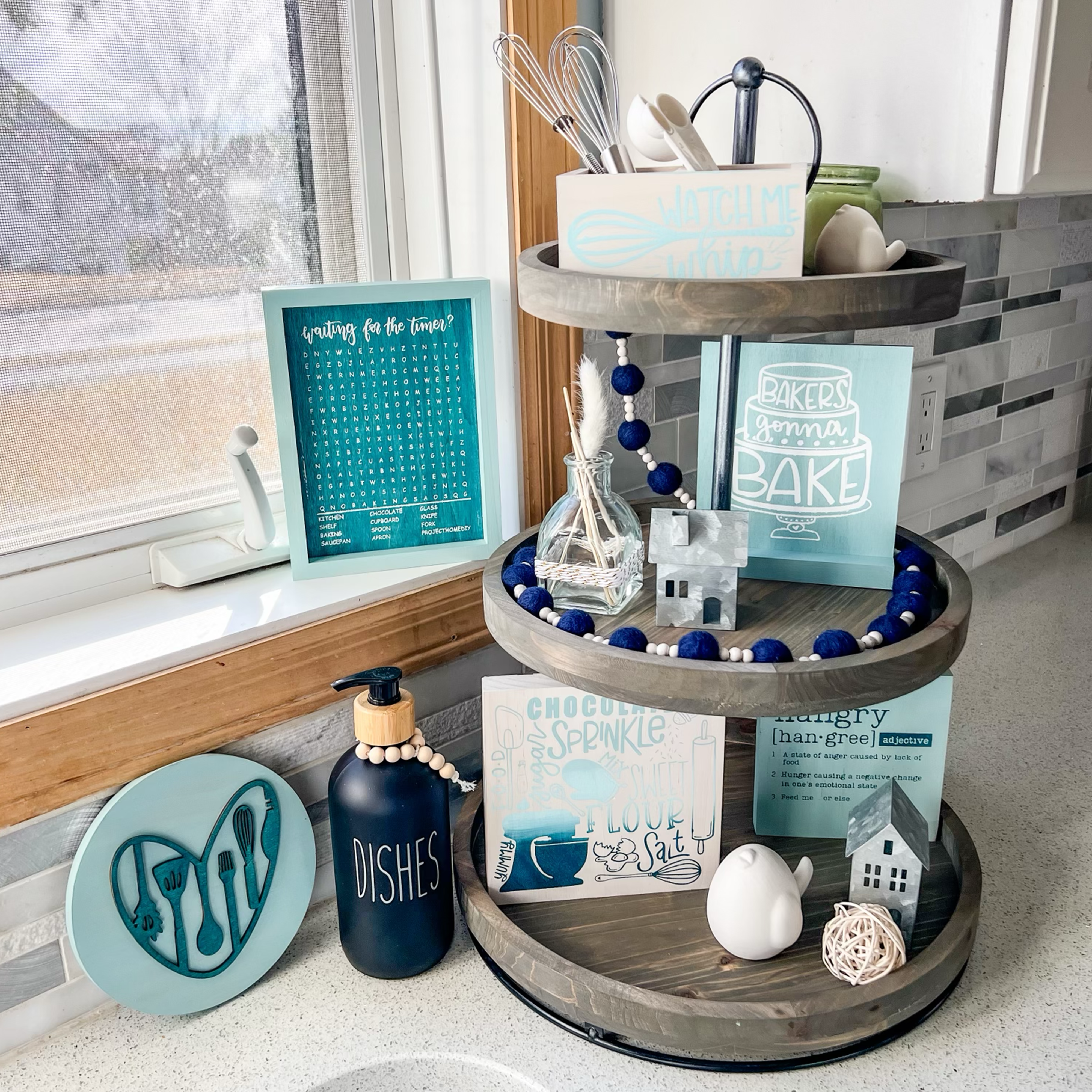 Two-tiered kitchen decor display with various baking-themed items and a word search by a sunny window.