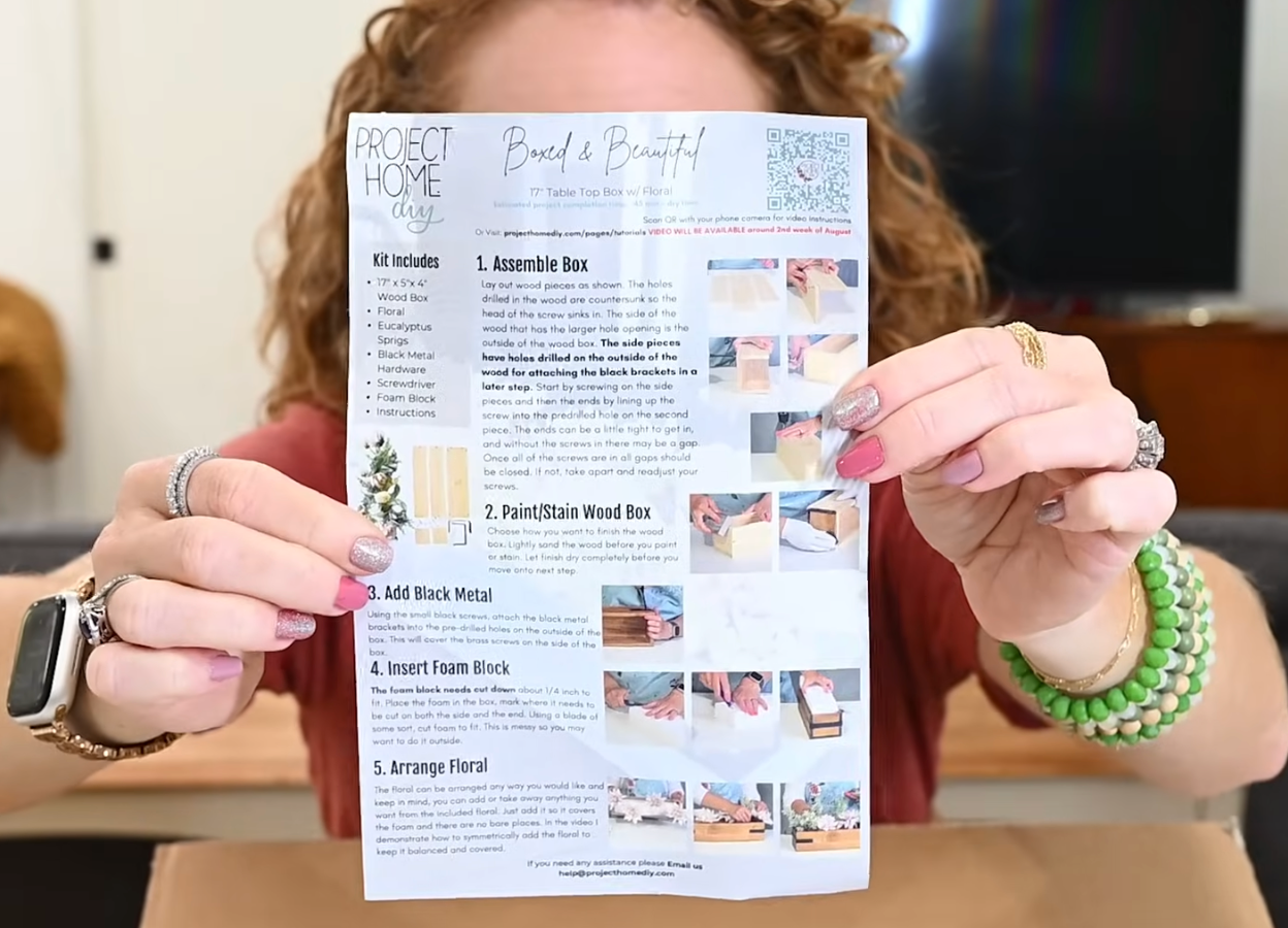 Person holding a 'Project Home DIY' instruction sheet for assembling a wood box and arranging floral decorations.