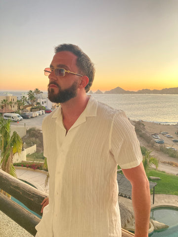 bearded man wearing gold and brown square sunglasses in front of sunset