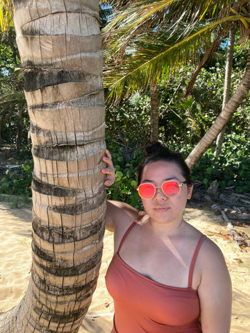 woman on tropical beach standing next to palm tree while wearing pink mirrored sunglasses