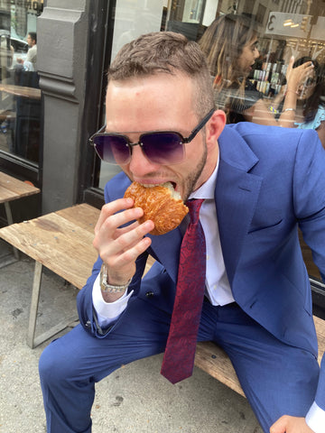 man wearing a suit and mirrored aviator sunglasses eating a croissant on a new york city street